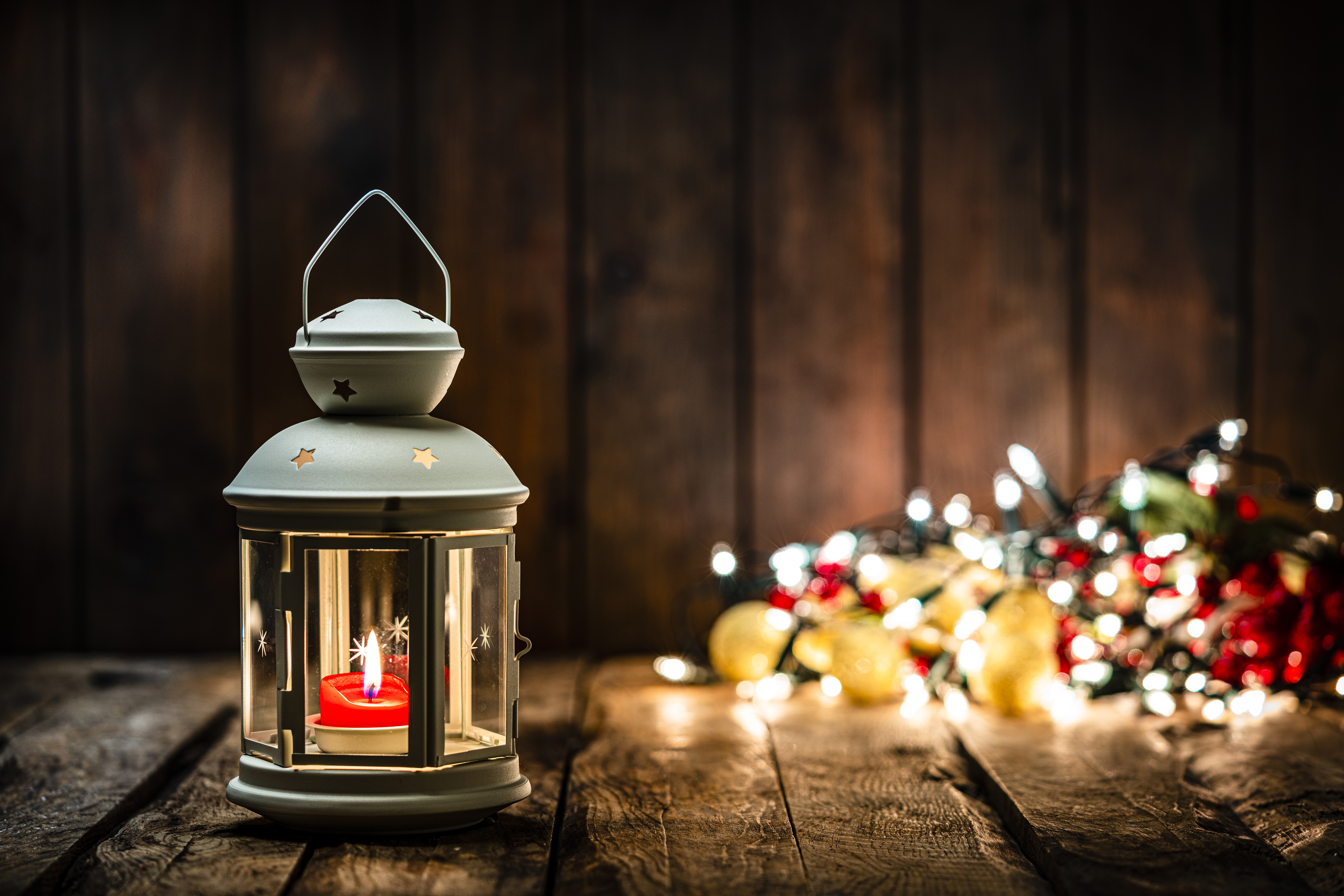 Christmas backgrounds: Christmas lantern on rustic wooden table. The composition is at the left of an horizontal frame leaving useful copy space for text and/or logo at the right. String light and Christmas decoration are out of focus at background. Predominant colors are yellow and brown. High resolution 42Mp studio digital capture taken with Sony A7rII and Sony FE 90mm f2.8 macro G OSS lens