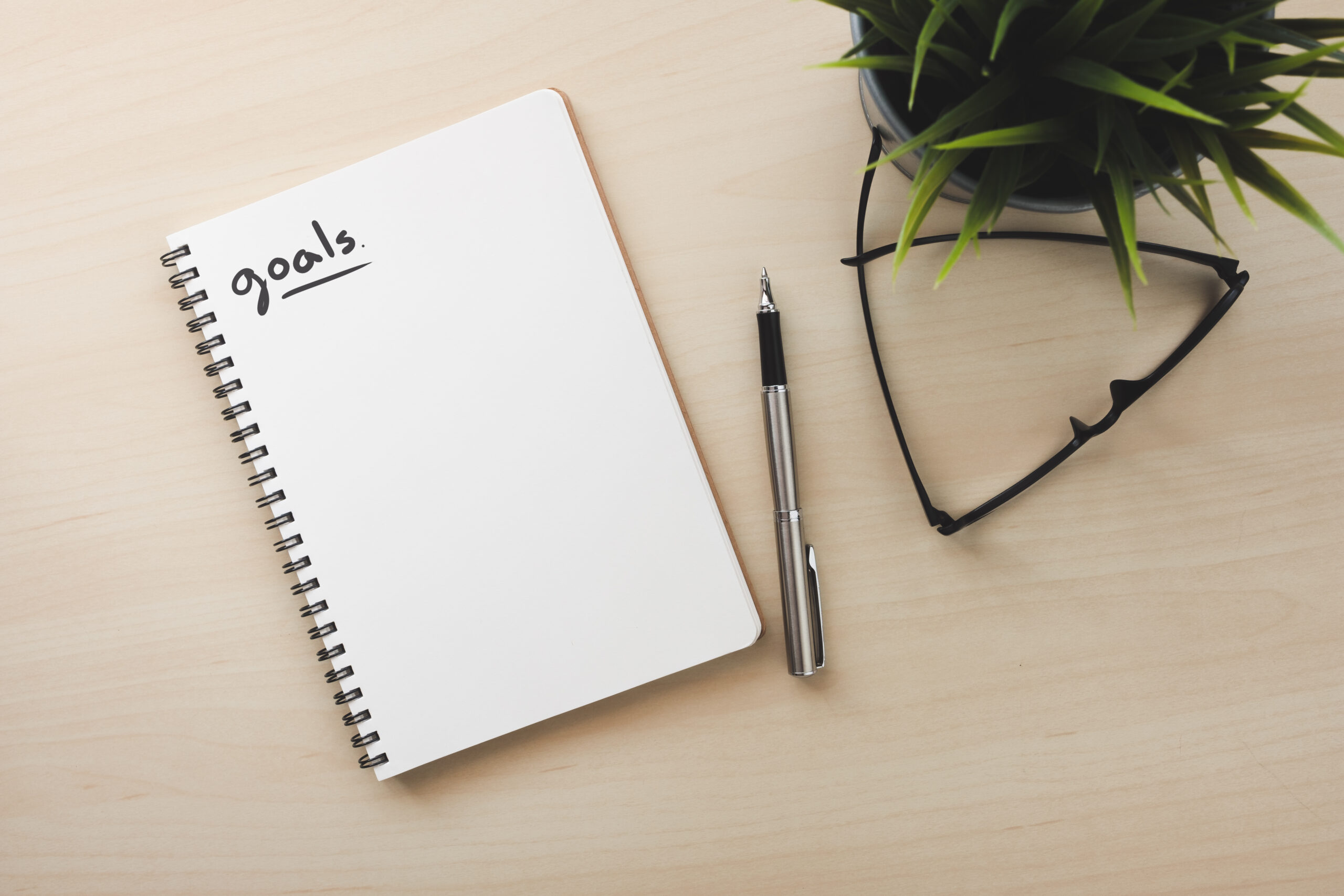 White notebook top view with small goals words on wood table close to pen and glasses