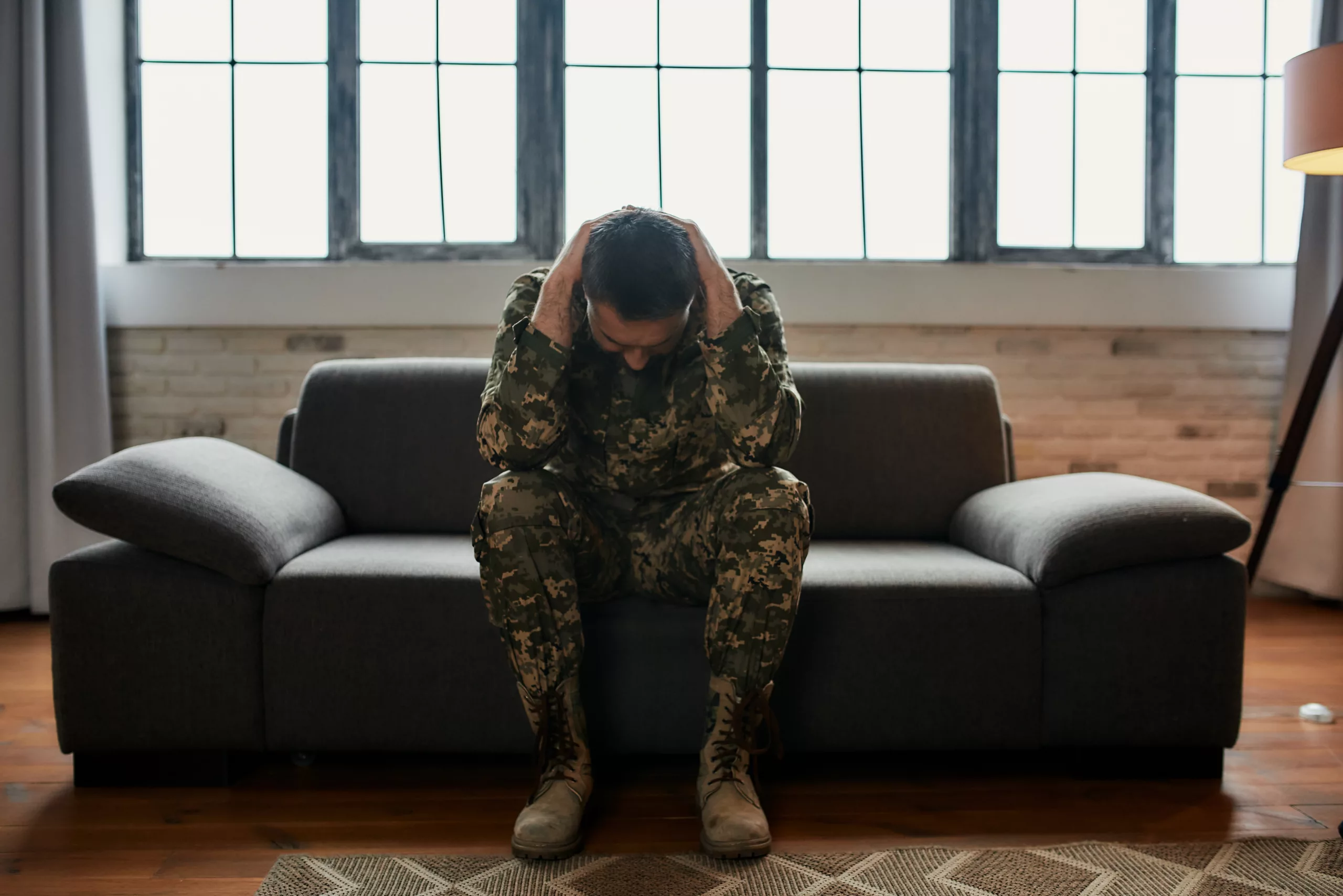 Full length shot of middle aged military man holding his head in pain and depression sitting on the couch. Soldier suffering from psychological trauma. PTSD concept. Front view. Horizontal shot