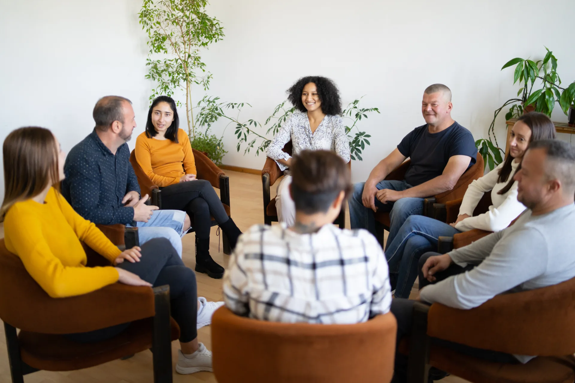 A group of mixed ethnic people sitting in a circle in comfortable chairs holding a support meeting, as they smile and chat with each other