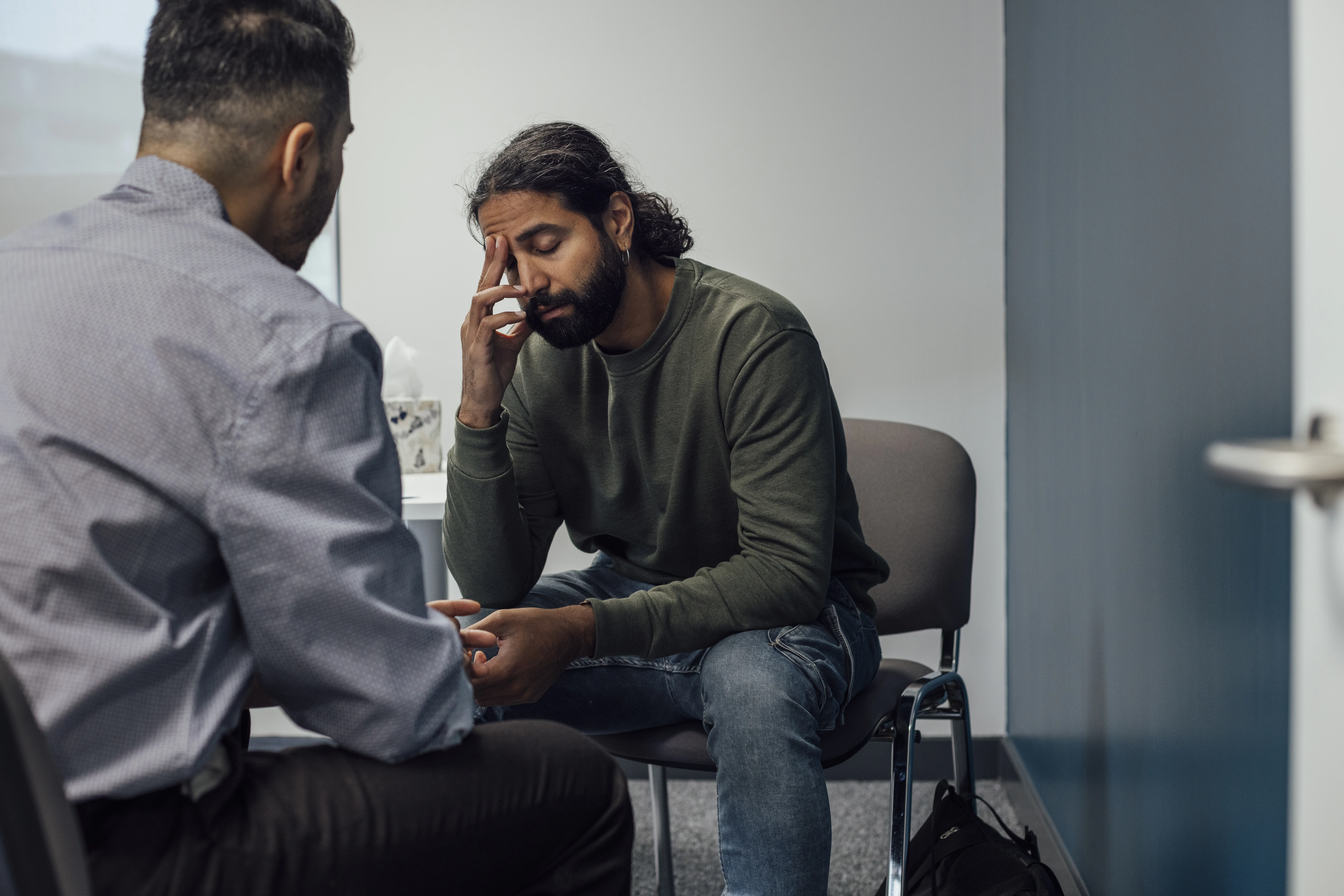 Over-the-shoulder shot of a doctor and man. The man looks sad and depressed after hearing news from the doctor. The man's eyes are closed and has one hand covering part of his face.